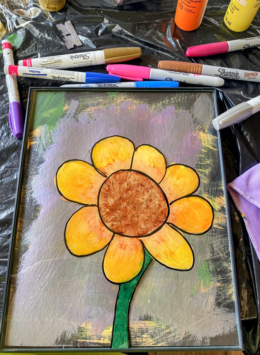 A bright sunflower painted on glass, resting on the work table among markers and paints